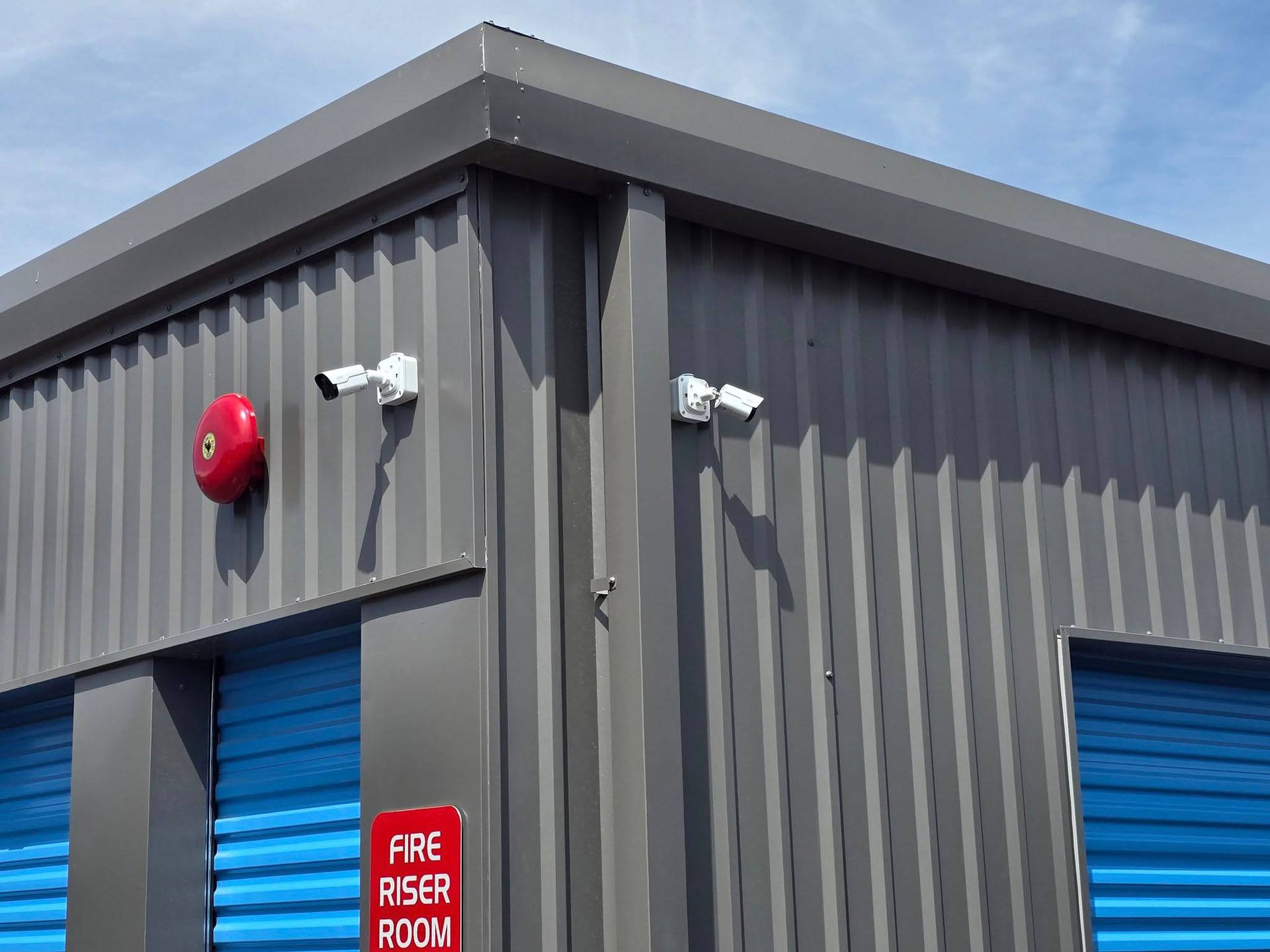 Gray metal building corner with cabling and two security cameras, a red fire alarm, a "Fire Riser Room" sign, and blue roll-up doors under a clear sky.